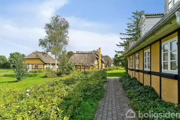 Gule, stråtækte huse står langs en stenbelagt sti i en grøn have. Store træer omgiver området under en blå himmel.