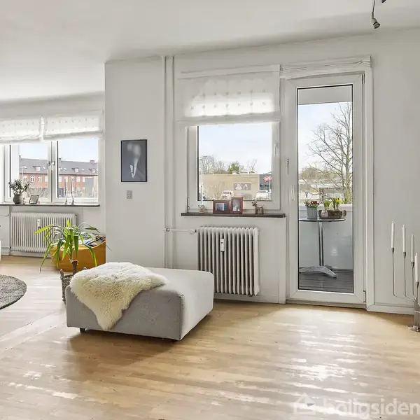 Stue med lys indretning har en stor sofa og træmøbler, vinduer med udsigt og en glasdør til altanen. Plante og dekorative genstande står rundt i rummet.