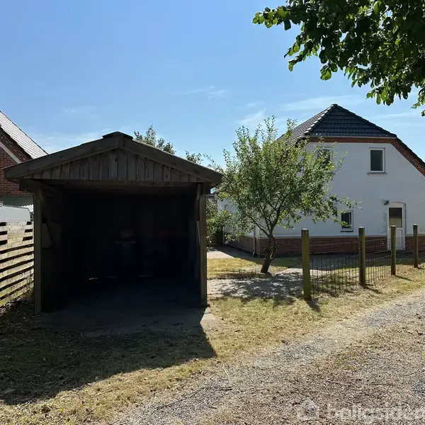 En trægarage står tom ved siden af en hvid murstensvilla. Villaen er omgivet af et metalhegn, træer og en grusvej. I baggrunden ses flere huse.