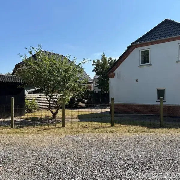 Et hvidt hus med mørkt tag står bag et lavt hegn. Til venstre ses et træ og en carport. I baggrunden er grønne buske og flere bygninger.