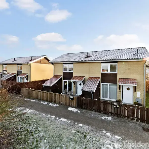 To rækkehuse med skråtag står langs en grusvej, omkranset af hegn og hække, under en blå himmel med spredte skyer. Snepletter ses på jorden.