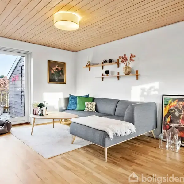 En grå sofa står i en lys stue med træloft og parketgulv. Ved siden af er en plakat, og flere planter står nær en glasdør med udsigt til en balkon.