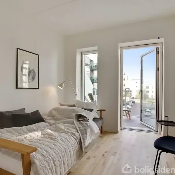 Sofa med tæppe og puder står i et lyst værelse. En åben dør leder til en balkon. Værelset har en moderne stol, en læselampe og en hylde på væggen.