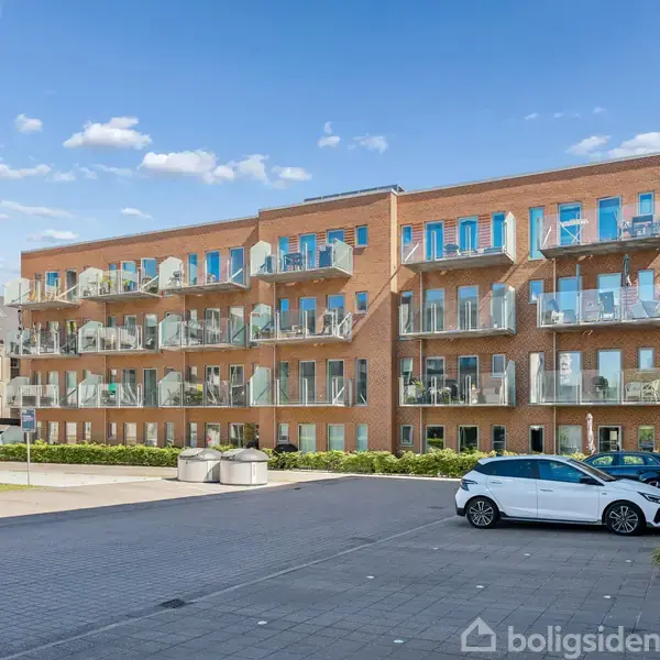 Bygning i røde mursten med mange altaner, stille på en asfalteret plads under blå himmel. Til venstre står et højt skilt med teksten "SOLBJERG KOLLEGIE", og en bil er parkeret i nærheden.