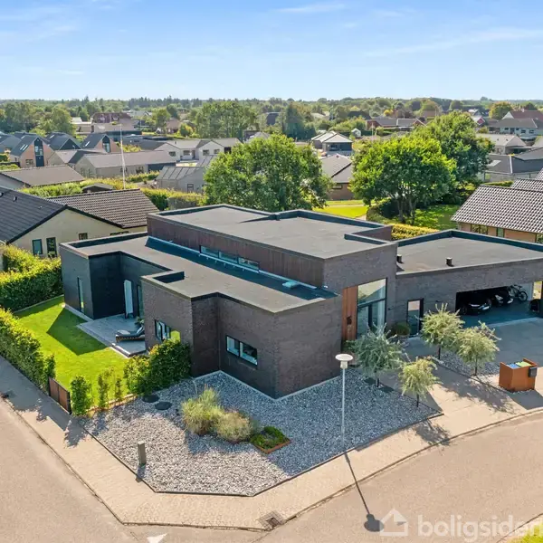 Et moderne, fladt-tagget hus ligger på et hjørne i et boligområde. Omgivet af en velplejet have med grønne buske. Nabohuse og træer ses i baggrunden.