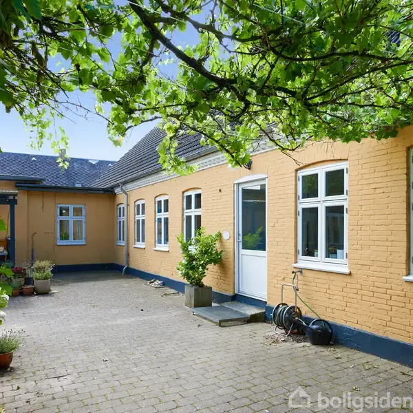 Hus med gule murstensvægge og hvide vinduer omgiver en brostensbelagt gårdhave. Klatreplanter hænger fra overhængende strukturer. Potteplanter og en haveslange står langs væggen.