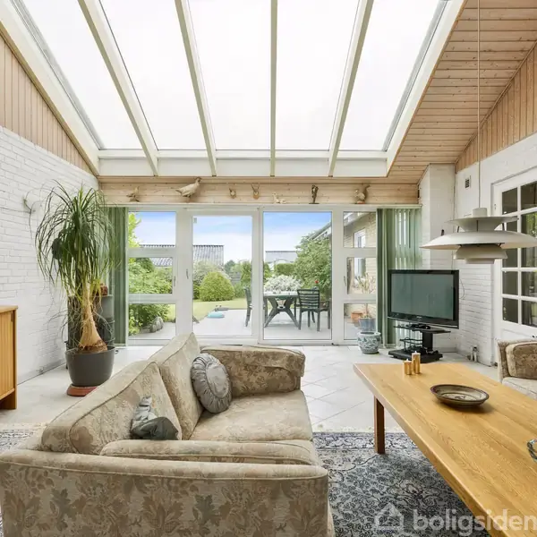 Sofa og sofabord står i en lys, moderne stue med glastag. TV'et står ved siden af en glasskydedør, der åbner til en terrasse med udsigt over en grøn have. En plakat med teksten ”JOURNEY” hænger på væggen.