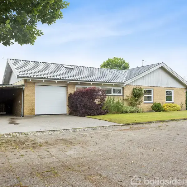 En gul murstensbungalow med grå tagsten står i et villakvarter. Huset har en garage og en anlagt forhave med buske. Flisebelagt indkørsel fører til garagen.