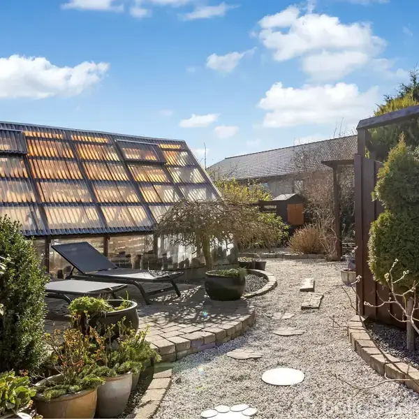 Liggestol ligger på en solrig terrasse omgivet af grønne planter og små træer i potter. Grussti med dekorative sten fører gennem haven. Velplejet og afslappende udendørsområde med blå himmel.