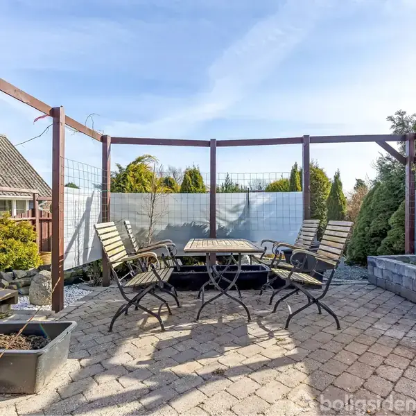 Fem havebordsstole står omkring et bord på en brolagt terrasse. Rundt om er der metaltrådshegn og buske, hvilket skaber en hyggelig og afskærmet atmosfære.