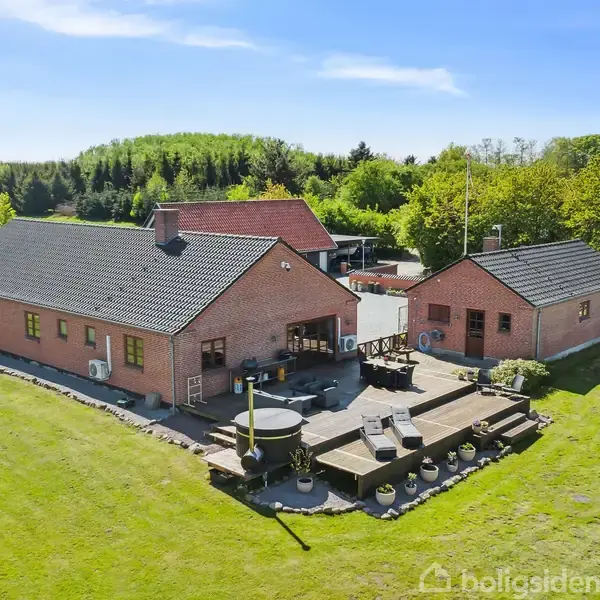Et rødstenshus med terrasse er omgivet af en grøn have. Der er havemøbler på terrassen og en rund swimmingpool i græsplænen. Skoven ses i baggrunden.