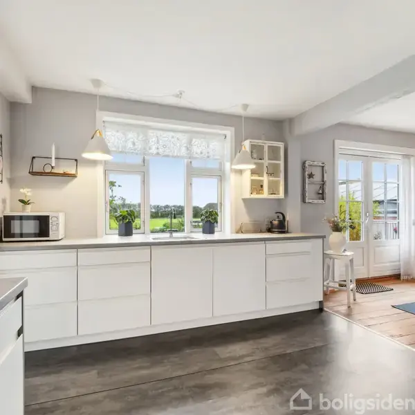 Køkken med hvidt skabsarrangement; vinduer viser en grøn udsigt. Hylder med dekorationer hænger på væggen. Tilstødende rum har stole og et tæppe. Gulvlampen lyser området op.