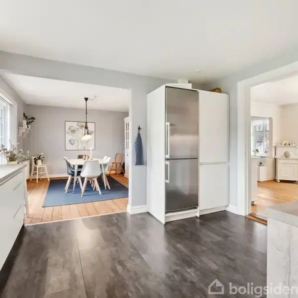 Køkken med hvide skabe og en stor sølvfarvet køleskab. Spisebord med seks stole står på et blåt tæppe i spisestuen. Lysegrå vægge og mørkt trægulv.