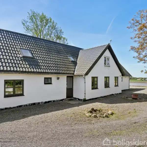 Hvid murstensbygning med sort tegltag står stille i en grusbelagt gårdsplads. Omkring huset er træer og en klar blå himmel i landlig omgivelser.
