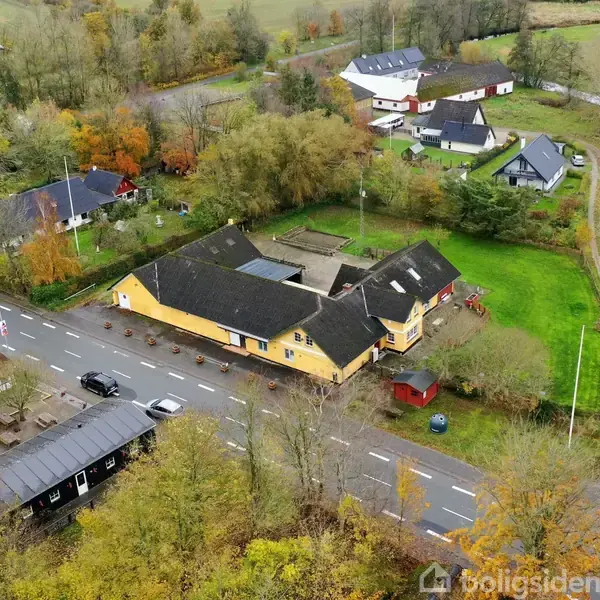 Et gult hus, omkranset af træer, ligger ved en vej med biler parkeret foran. Omgivelserne består af flere bygninger og grønne, skovklædte områder.