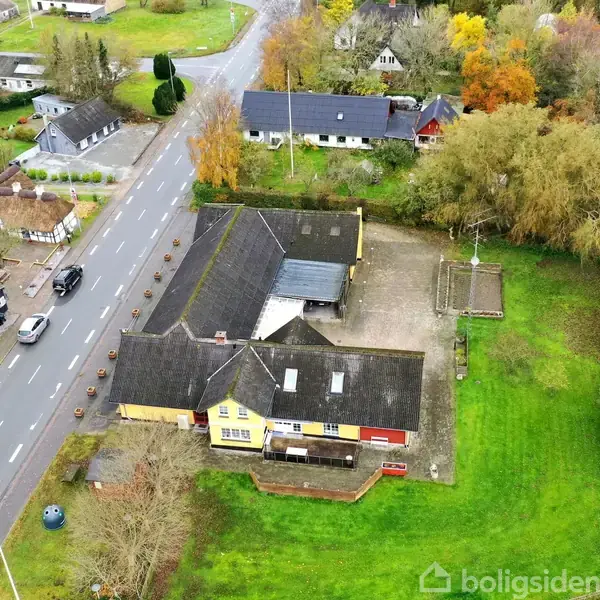 En gul bygning ligger ved en vej med biler, omgivet af grønne træer og græsområder. Flere mindre huse ses i baggrunden.