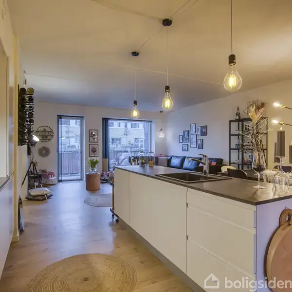 Et moderne køkken-alrum med hvide skabe og en stor køkkenø i forgrund. Rummet er lyst med hængende lamper, trægulv og store vinduer. Møbler inkluderer sofa og spisebord.