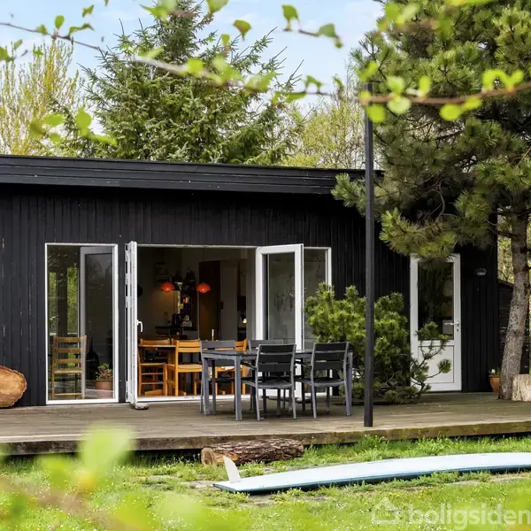 Sort træhus med åbne døre, der fører til en terrasse med havemøbler. Omgivet af grønne træer og en græsplæne med et surfbræt.
