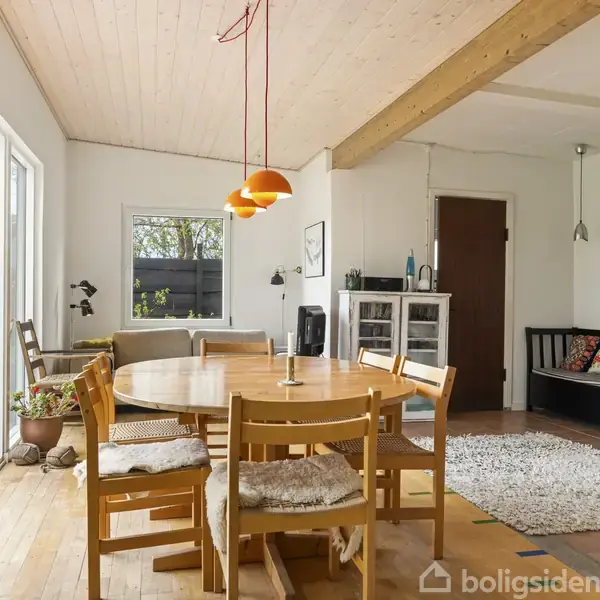 Rundt spisebord med træstole står under orange lamper i en lys stue. En dør fører ud til terrasse, og sofa står nær vindue. Tæppe på gulvet.
