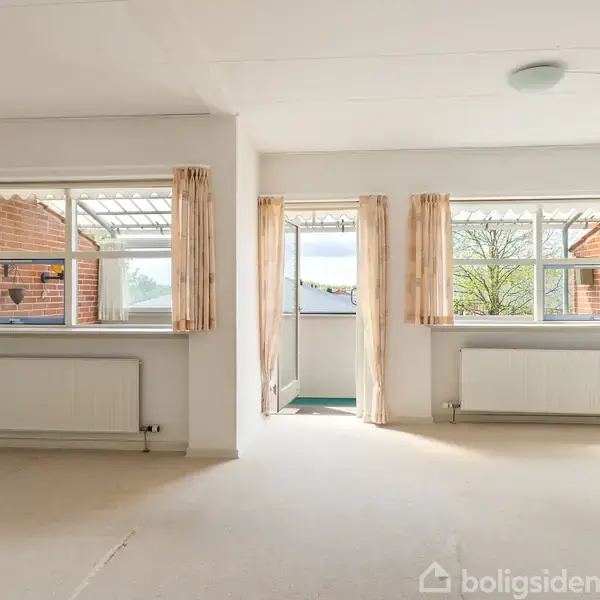 Et lyst værelse med to vinduer og en glasdør til en balkon. Rummet har hvide vægge og gulvtæppe; radiatorkasser under vinduerne. Gardiner hænger ved åbningerne.
