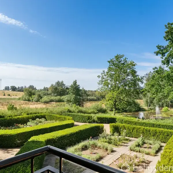 En velplejet have med hække arrangeret i geometriske mønstre, set fra en balkon i et hus. I baggrunden ses træer og en lille dam i naturligt landskab.