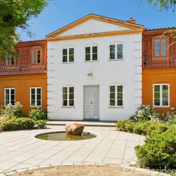 En hvid og orange villa med symmetrisk facade står i en frodig have. Foran huset er en rund terrasse med en lille vandinstallation og en stor sten i midten.