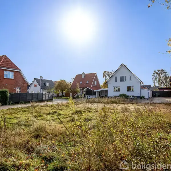 Huse står i solskin, omkranset af græsmark. Træer flankerer grunden, og den klare himmel danner en lys baggrund.