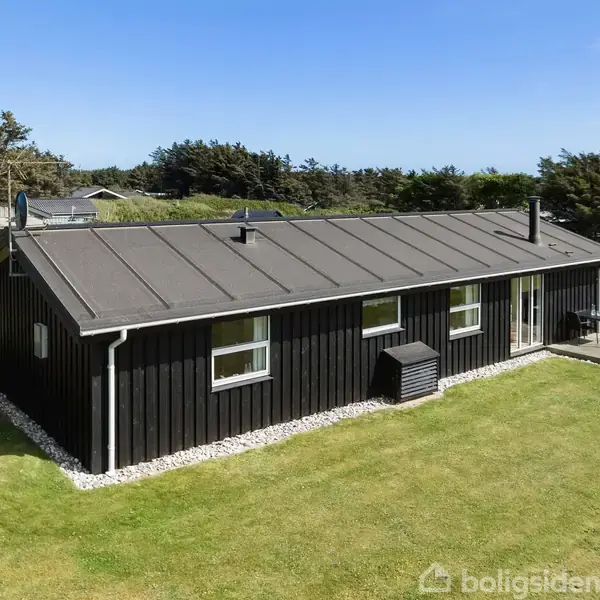 Et sort træhus står stille på en velholdt græsplæne. Det har store vinduer og en lille terrasse. Omkring huset er der grønt område med buske og træer under en blå himmel.