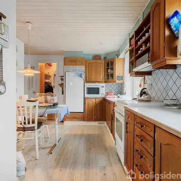 Et køkken med træskabe og hvidevarer langs væggen. En spiseplads med stole og et bord er synlig i baggrunden. Lyse trægulve og hvide loftpaneler kompletterer rummet.