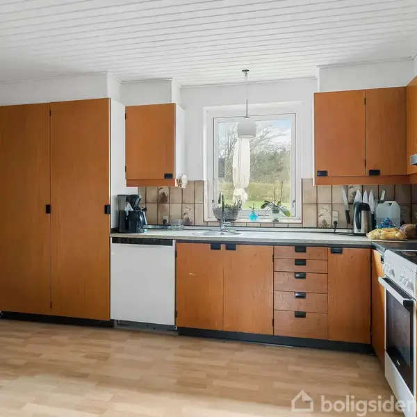 Køkken med brune skabe og hvide vægge, indeholder køleskab med magneter, ovn, og opvaskemaskine. Et rundt bord står i venstre hjørne. Vindue giver udsigt til grøn natur.