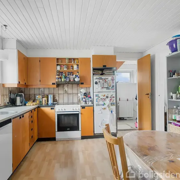 Køkken med lyse trægulve, træfarvede skabe og hvidt loft. Køkkenbordet rummer en kaffemaskine, frugt og opvaskemaskine. Et spisebord med stole står i forgrunden. En åben reol viser porcelæn og bøger.