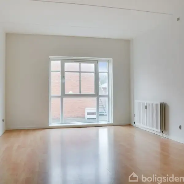 En tom stue står stille med trægulv, et stort vindue mod en murstensbygning og en radiator på højre væg. Rummet fremstår lyst og minimalistisk.