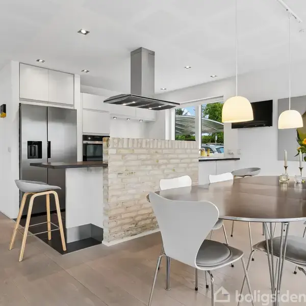 Køkken med moderne indretning: Der er en rund spisebord med seks stole, og en ø med barstole. Rummet er lyst og stilrent med lyse gulve og minimalistisk design.