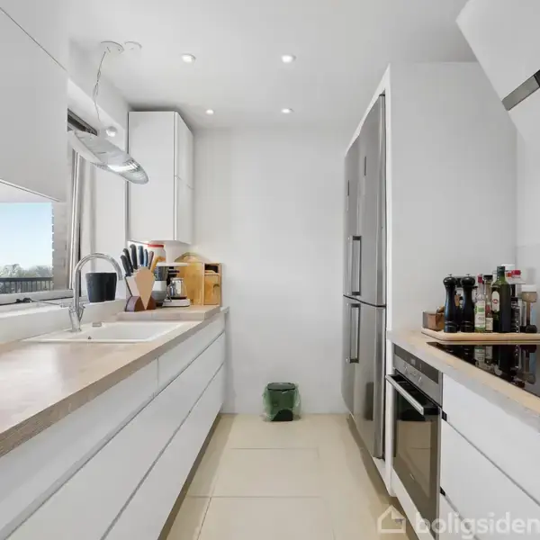 Et moderne køkken med hvidt inventar, to store køkkenborde og en stålvask. Der er en stor grå køleskab-fryser og indbygget ovn. Rummet er lyst med vindue til venstre.