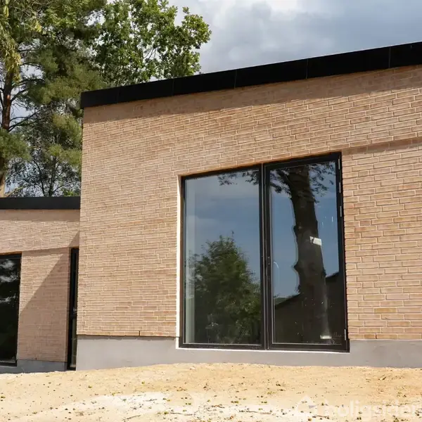 Et moderne hus med store glaspartier står i en lys byggeområde omkranset af træer. Den lyse murstensfacade skinner i solen og reflekterer omgivelserne i vinduerne.