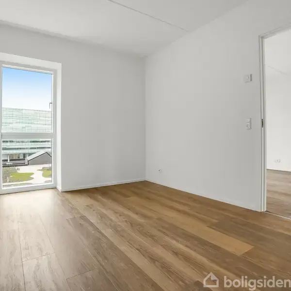 Et rum med trægulv og hvide vægge har en døråbning til et tilstødende værelse. En dør i baggrunden åbner til en balkon med udsigt over moderne bygninger.
