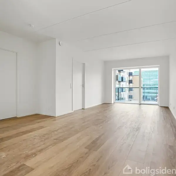 Et tomt rum med trægulv åbner ud mod en balkon gennem en stor glasdør. Baggrunden viser moderne lejlighedsbygninger. Væggene er hvide, og lyset er naturligt.