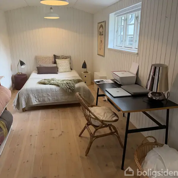 Seng med puder og plaid står i et lyst soveværelse. Til højre et skrivebord med bærbar, printer og lampe. Værelset har trævægge og et vindue, der giver naturligt lys.