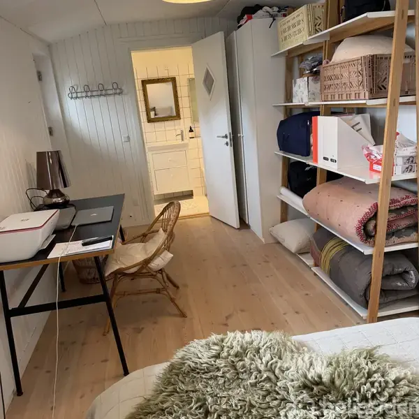 En arbejdsplads med skrivebord, bærbar computer og printer; omgivet af en stol. Åben dør fører til et badeværelse med vask. Til højre er der hylder med tæpper og kasser.