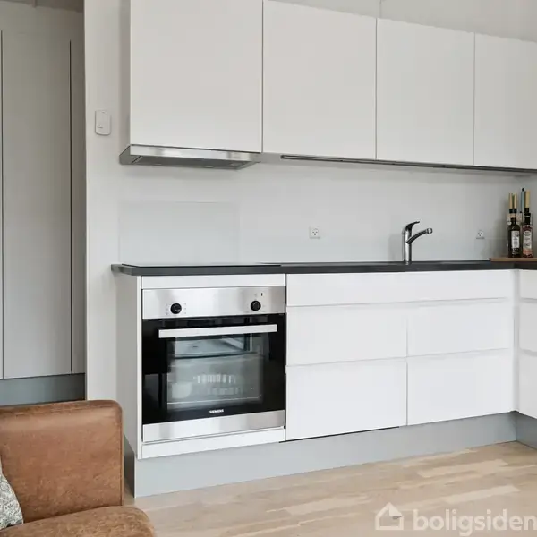 Hvidt køkken med indbygget ovn i et hjørne. Køkkenet har glatte skabe, sort bordplade, og flere flasker står ved vasken. En brun sofa ses i forgrunden.