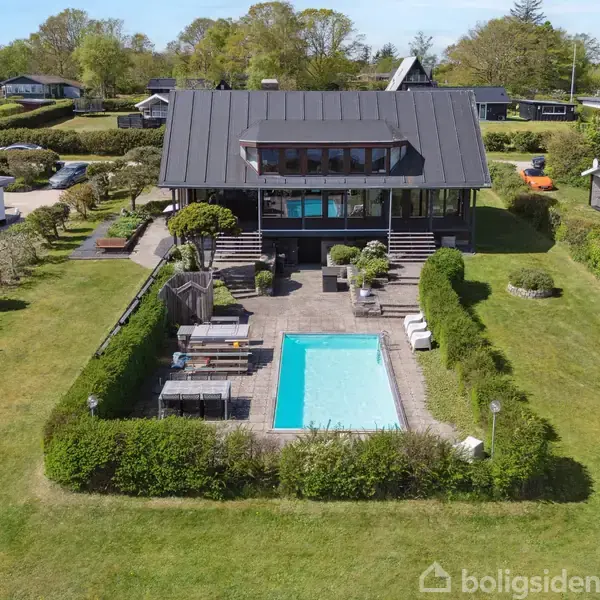 Et moderne hus med terrasse ligger i en grøn have med en swimmingpool. Andre huse ses i baggrunden, omgivet af grønne træer og buske.