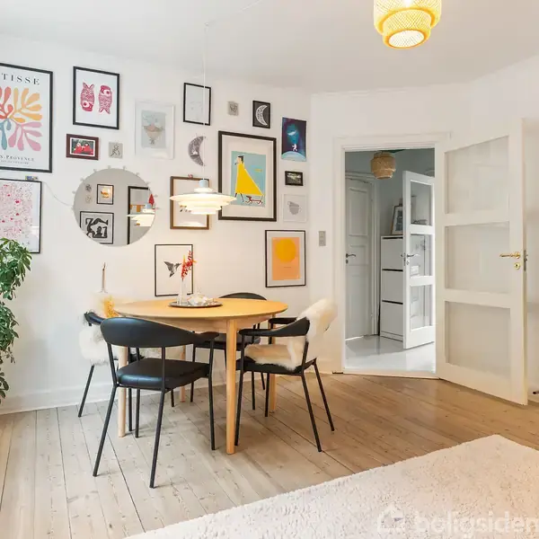 Rundt spisebord med fire stole står i en stue med lyse trægulve. Væggene er dekoreret med forskellige kunstværker. En stor plante står i et hjørne, og et tv står til højre.
