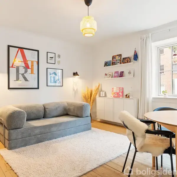 Grå sofa står i stue med kunst på væggen. Rundt bord med stole ved vindue, hylder med bøger og planter. Trægulv og hvidt tæppe danner hyggelig atmosfære.