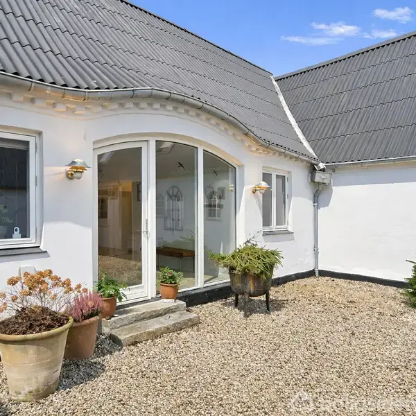 Et hvidt hus med en bueformet indgang, omgivet af grusbelagt gårdsplads og potteplanter. Store vinduer og en skrå tagflade sætter rammerne under en blå himmel.