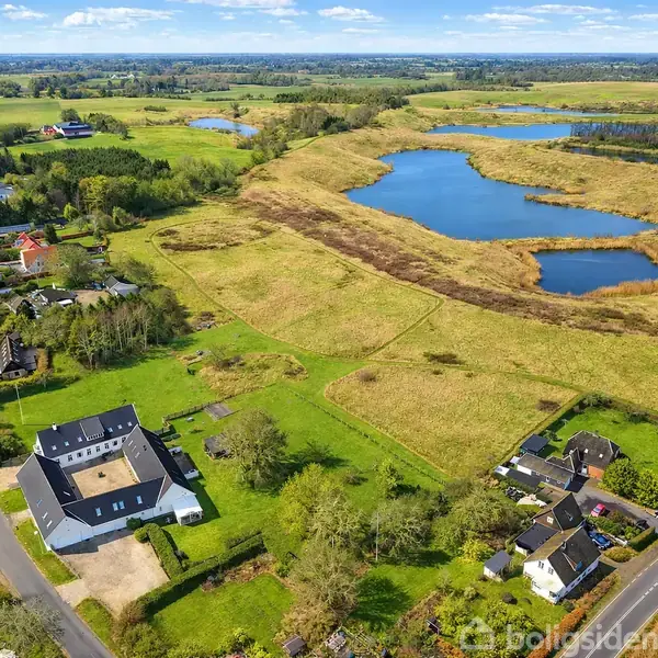 Et landskab med huse ved en landevej; grønne marker omgiver dem, og der er en stor sø i baggrunden. Himlen er blå med spredte skyer.