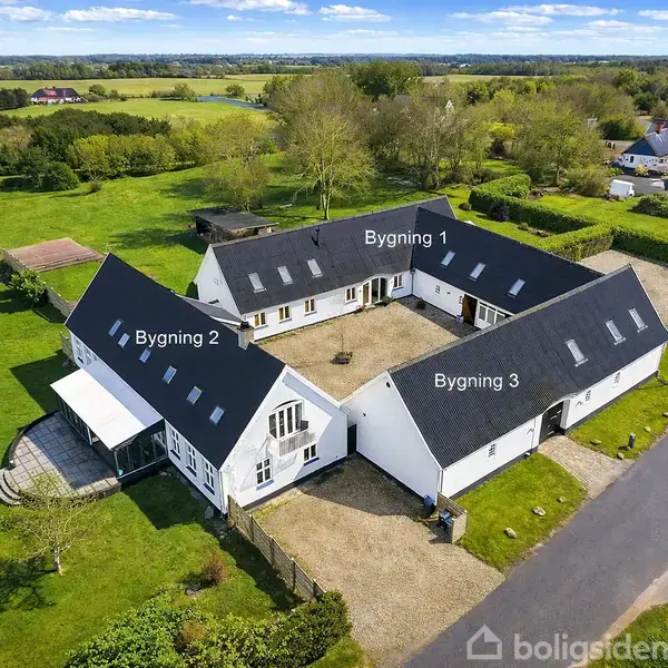Tre hvidmalede bygninger, mærket Bygning 1, Bygning 2, og Bygning 3, danner en gårdsplads på landet. Omgivet af grønne marker og en vej.