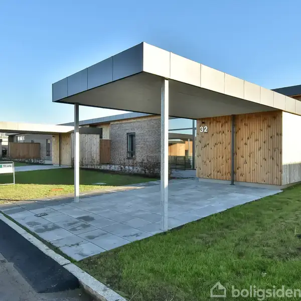 En moderne, lavtliggende murstensbygning med fladt tag har en carport nummereret "32" og er omgivet af grønne områder og en brolagt indkørsel. Det ligger i et boligområde.