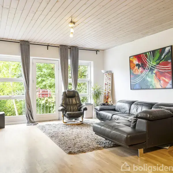 Stue med sort lædersofa og lænestol vender mod store vinduer med udsigt til have. Farverigt abstrakt maleri hænger på væggen. Trægulv og gulvtæppe skaber en hyggelig atmosfære.