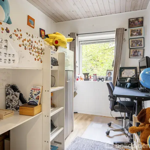 Et børneværelse med reol fyldt med bøger og pynt. Et skrivebord med stol står ved et vindue. Bamse og klode er på gulvet. Farverige vægdekorationer ses rundt omkring.