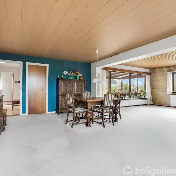 Et rummeligt opholdsrum med træloft og hvide vægge. Et spisebord med stole står ved siden af en gammel skænk. Store vinduer giver udsigt til en grøn have udenfor.